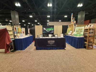 UConn booth with a central table displaying informational materials, flanked by blue‑skirted display tables on both sides inside a large convention hall.