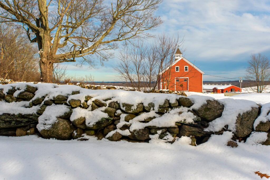 Snow‑covered stone wall in front of a red barn and outbuildings under a winter sky.