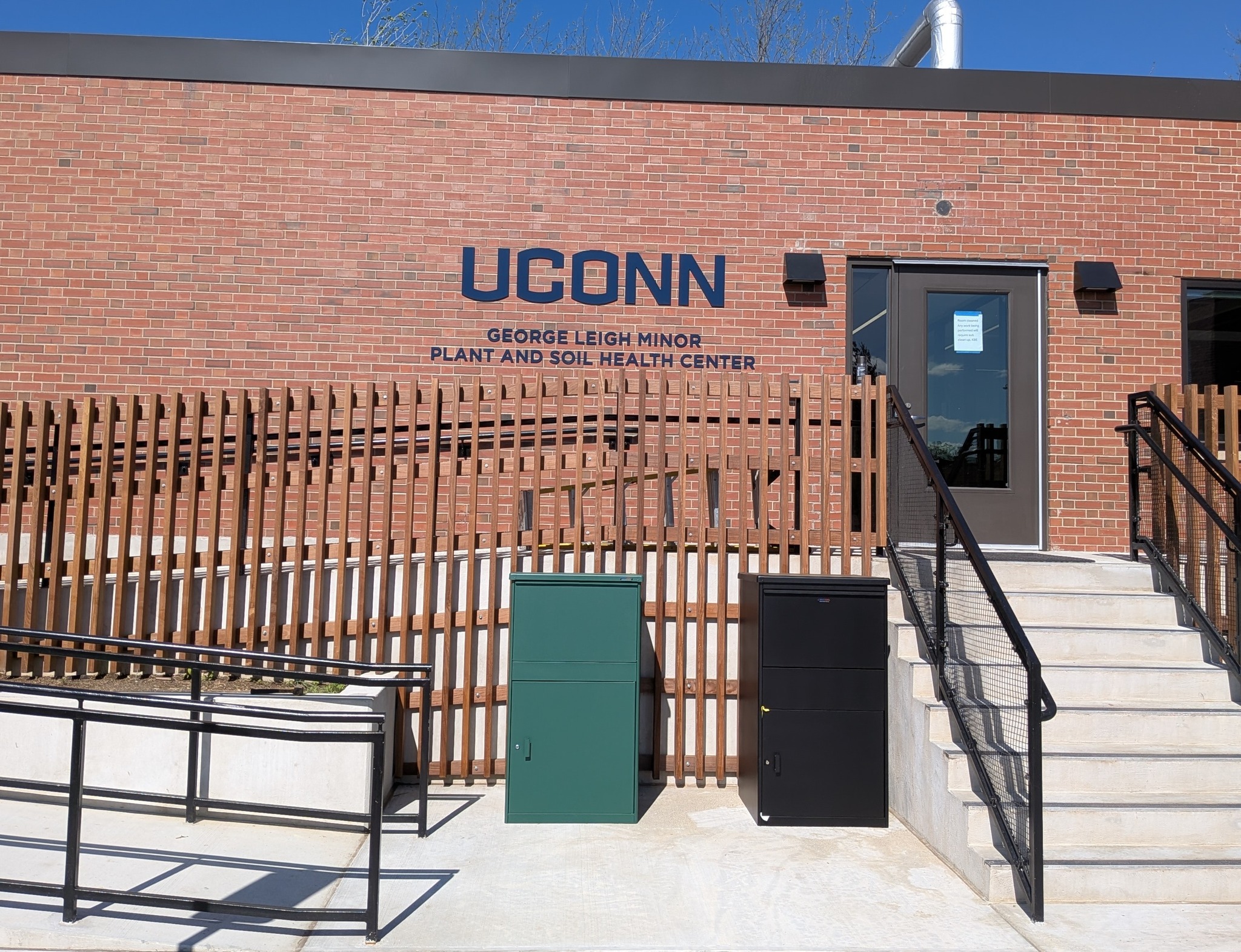 Brick building exterior with a sign reading “UConn George Leigh Minor Plant and Soil Health Center,” featuring a ramp, stairs, metal railings, and utility cabinets in front of a wooden slat fence.