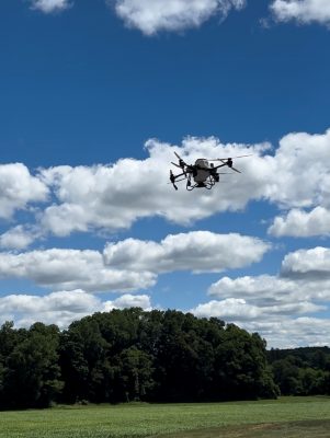 Drone flying in a blue sky with scattered white clouds above a grassy field and a line of trees in the background.