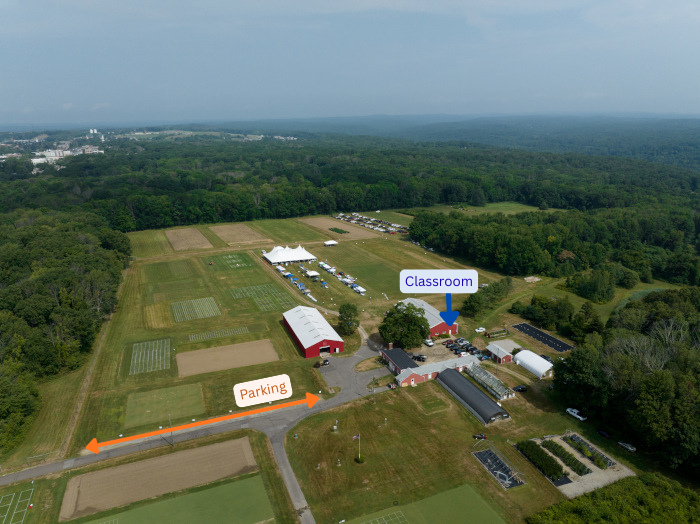Aerial view of a research farm with agricultural test plots, several farm buildings, and surrounding forest. Orange arrows label the route to “Parking,” and a blue marker identifies a building labeled “Classroom.”