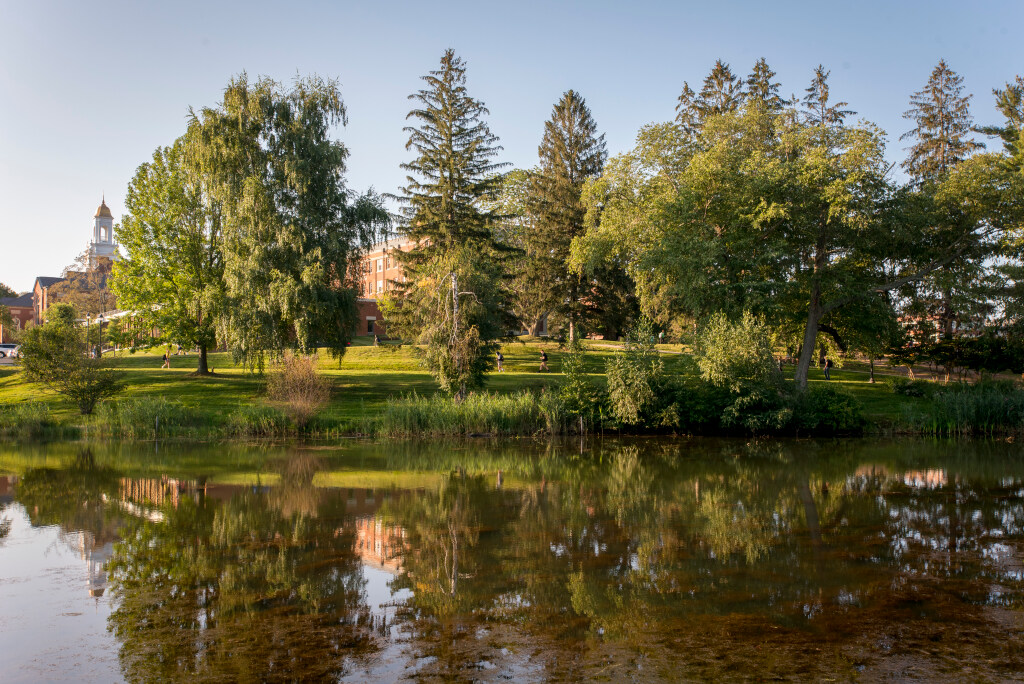 Campus pond reflecting trees and nearby buildings on a sunny day, with a grassy lawn and walking paths along the shoreline.